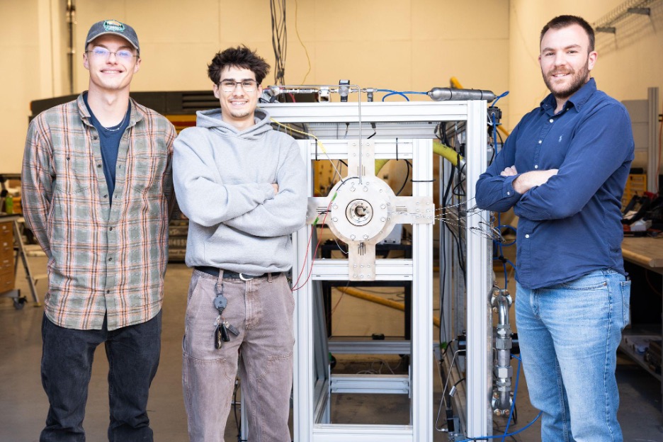 photo: Left to right: Undergraduate Mechanical Engineering students Micah Wolbrecht and Brendan Canales stand in front of the Rotating Detonation Engine Materials Testbed with Assistant Professor George Koutsakis.